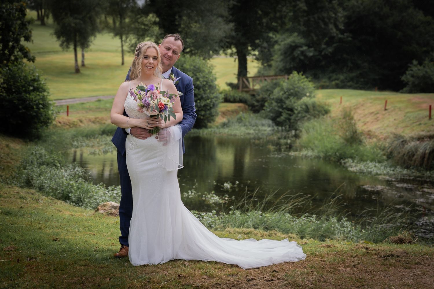 Wedding couple embracing on wooden bridge with lush summer landscape and tranquil pond in background.