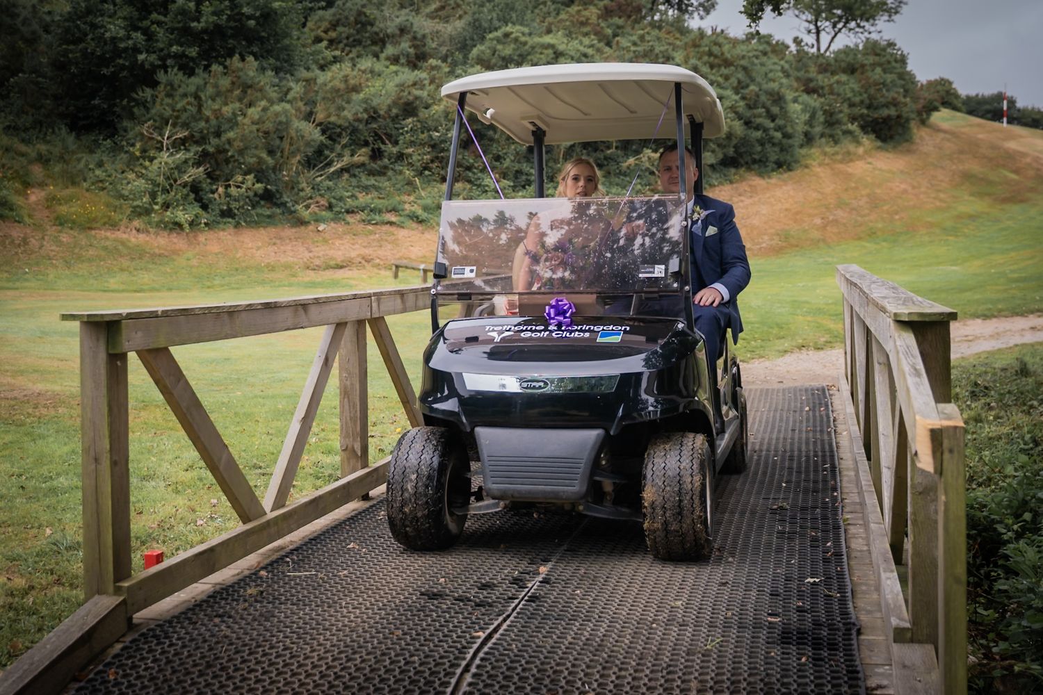A golf cart drives across a wooden bridge on a golf course path during a cloudy day.