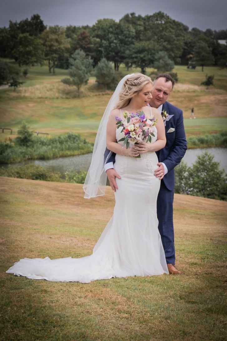 Bride and groom share a romantic embrace on a grassy hillside during their outdoor wedding photoshoot.