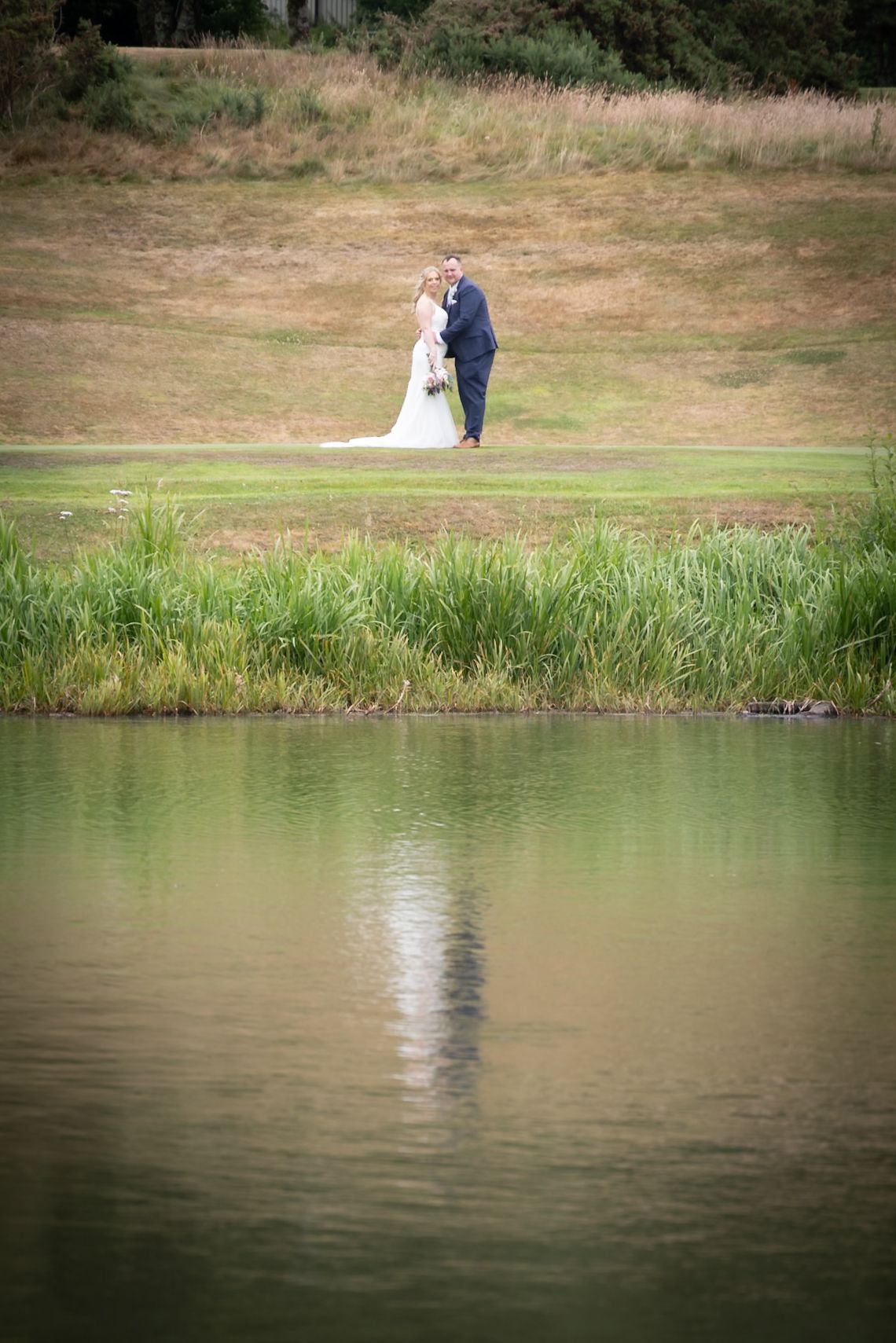A romantic wedding couple stands at the edge of a tranquil pond surrounded by green grass and reeds.