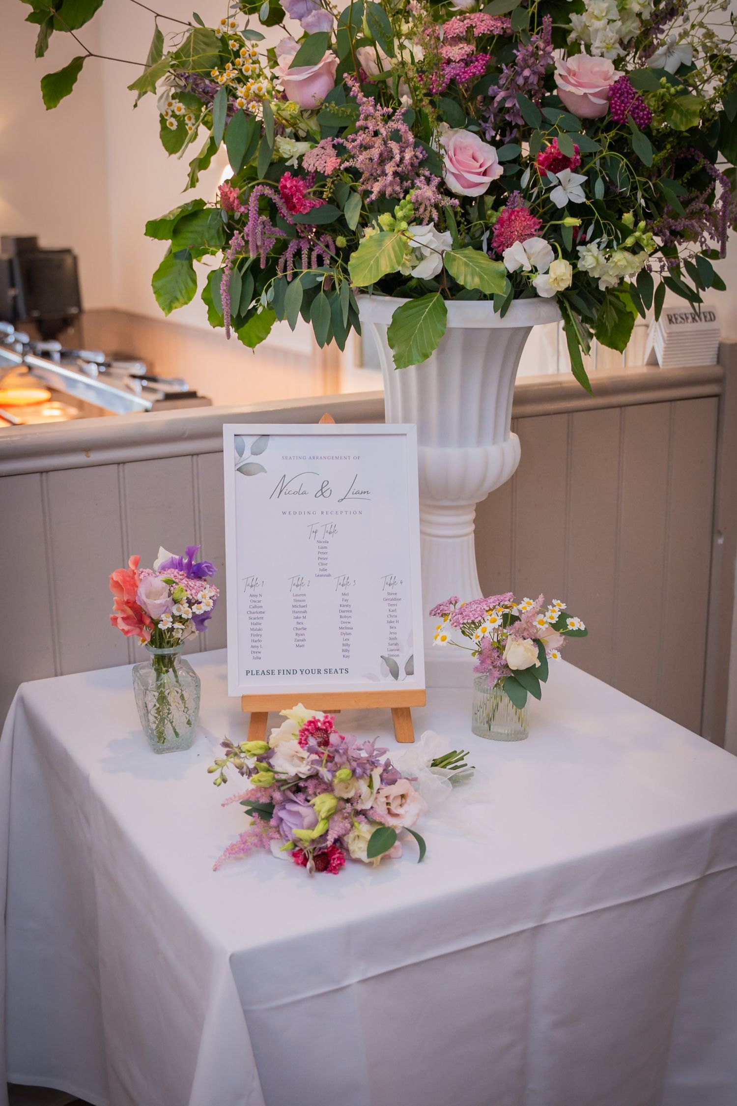 Elegant wedding reception table with white tablecloth, floral centerpiece in white urn vase, and framed menu card on easel.