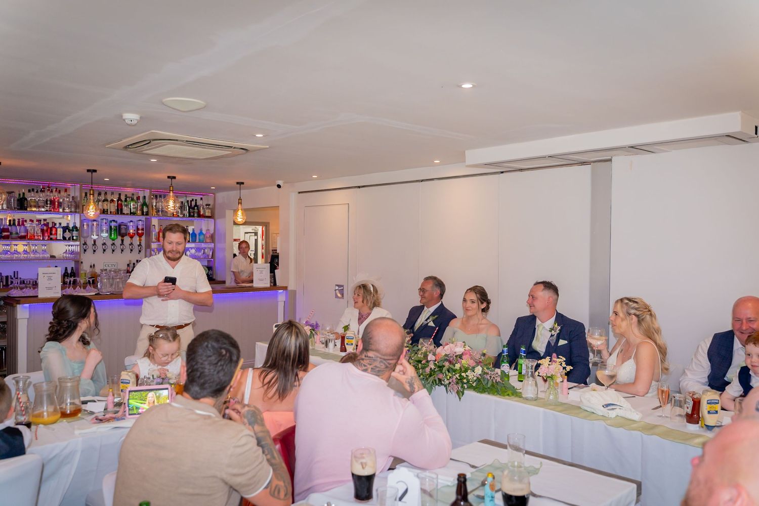 Wedding reception guests seated at decorated tables in a venue with purple ambient lighting and white floral centerpieces.