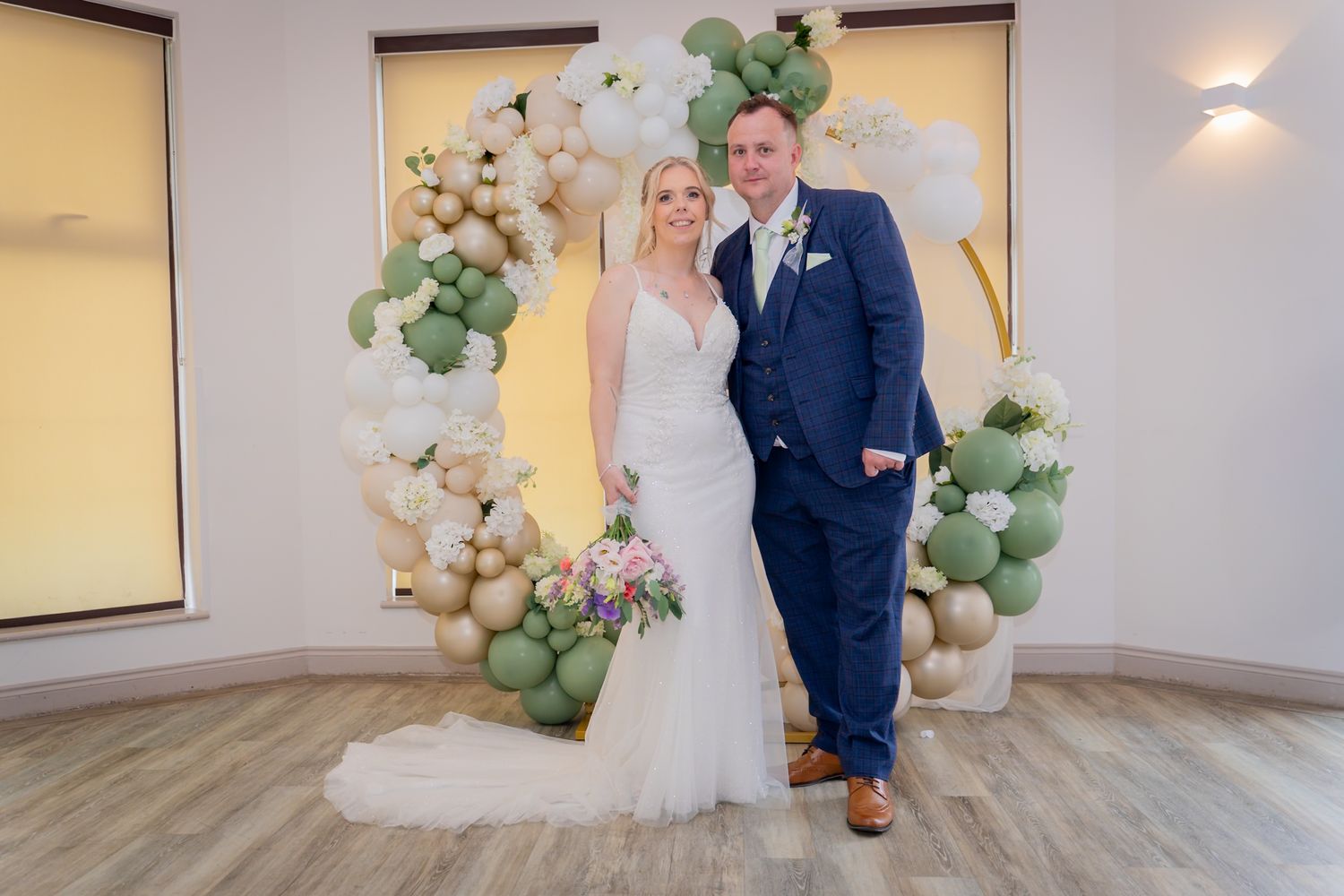 Wedding couple poses in front of elegant balloon arch and floral decorations at reception venue.
