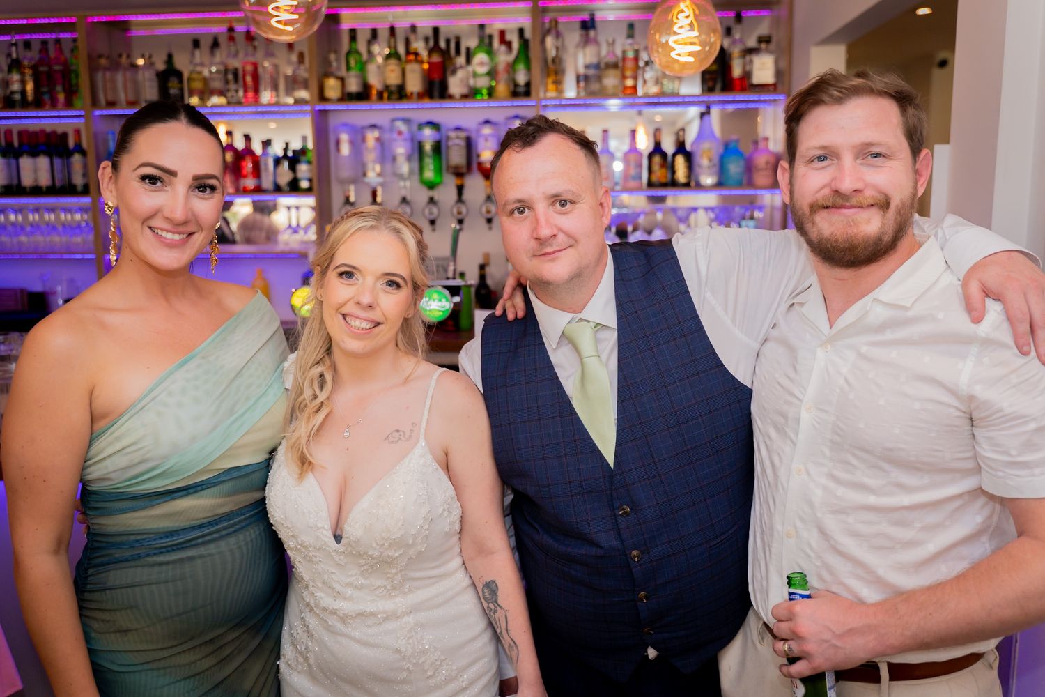Group photo of wedding party members smiling together at a reception with bar and purple lighting in background.