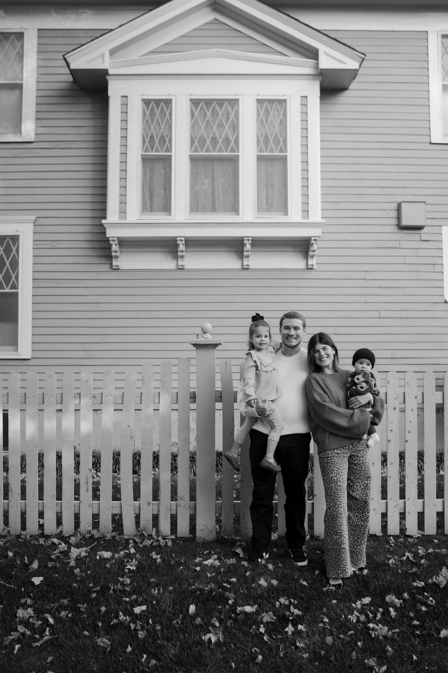 Family enjoying autumn day together by white picket fence and gray house.