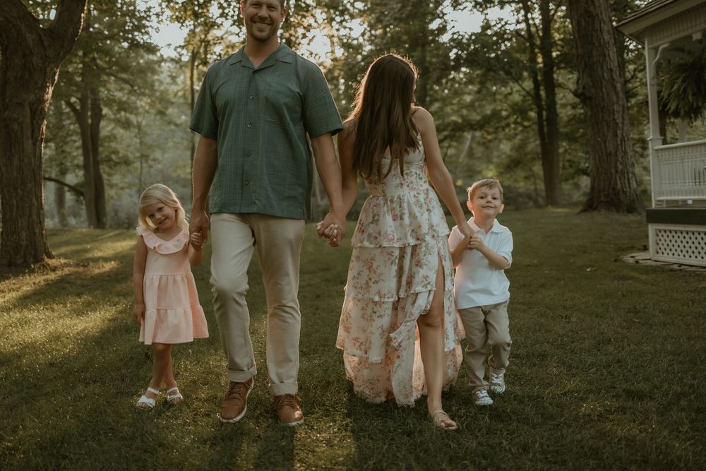 A family walks together in a sunlit forest setting wearing coordinated outfits in earth tones and floral patterns.