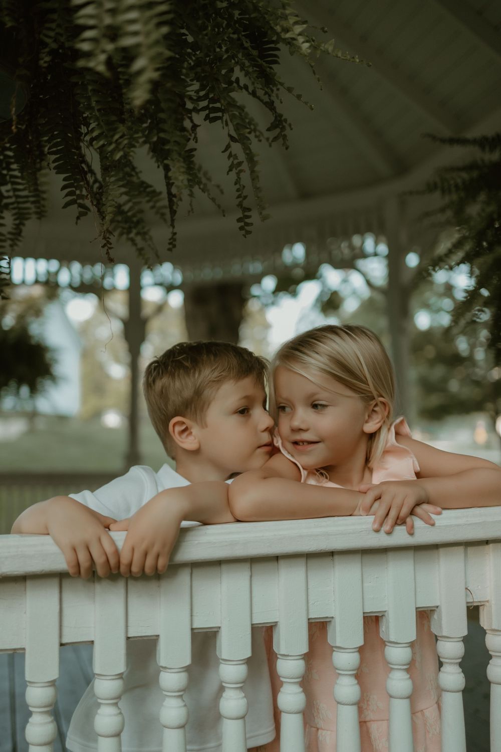 Two children lean against a white wooden railing while sharing a sweet moment together outdoors.
