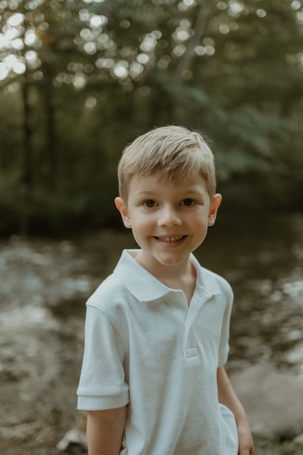 A young child in a white polo shirt smiles outdoors with bokeh lighting and trees in the background.