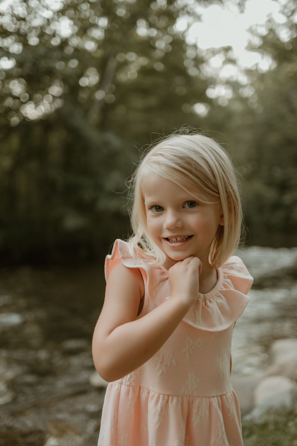 A young child in a peach-colored dress smiles playfully while holding their face.