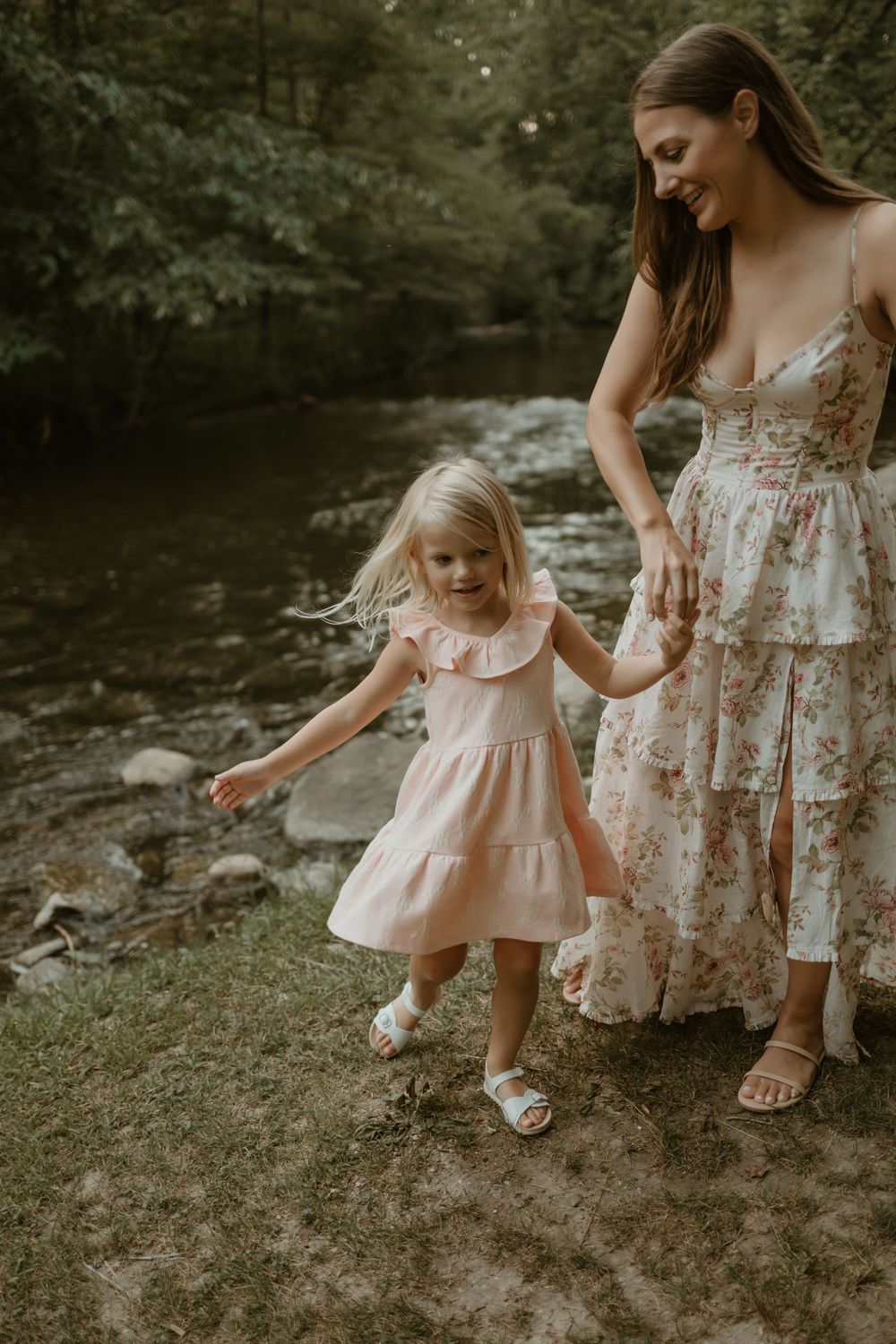A mother and child in coordinating floral dresses explore a grassy area near a stream.