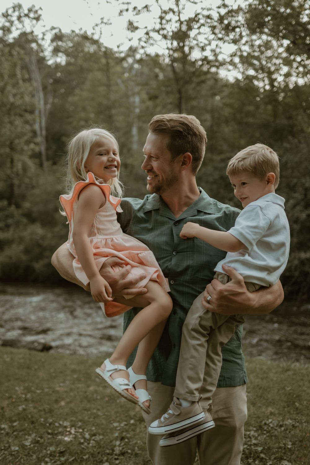 A parent holds two young children outdoors in a natural setting during golden hour.