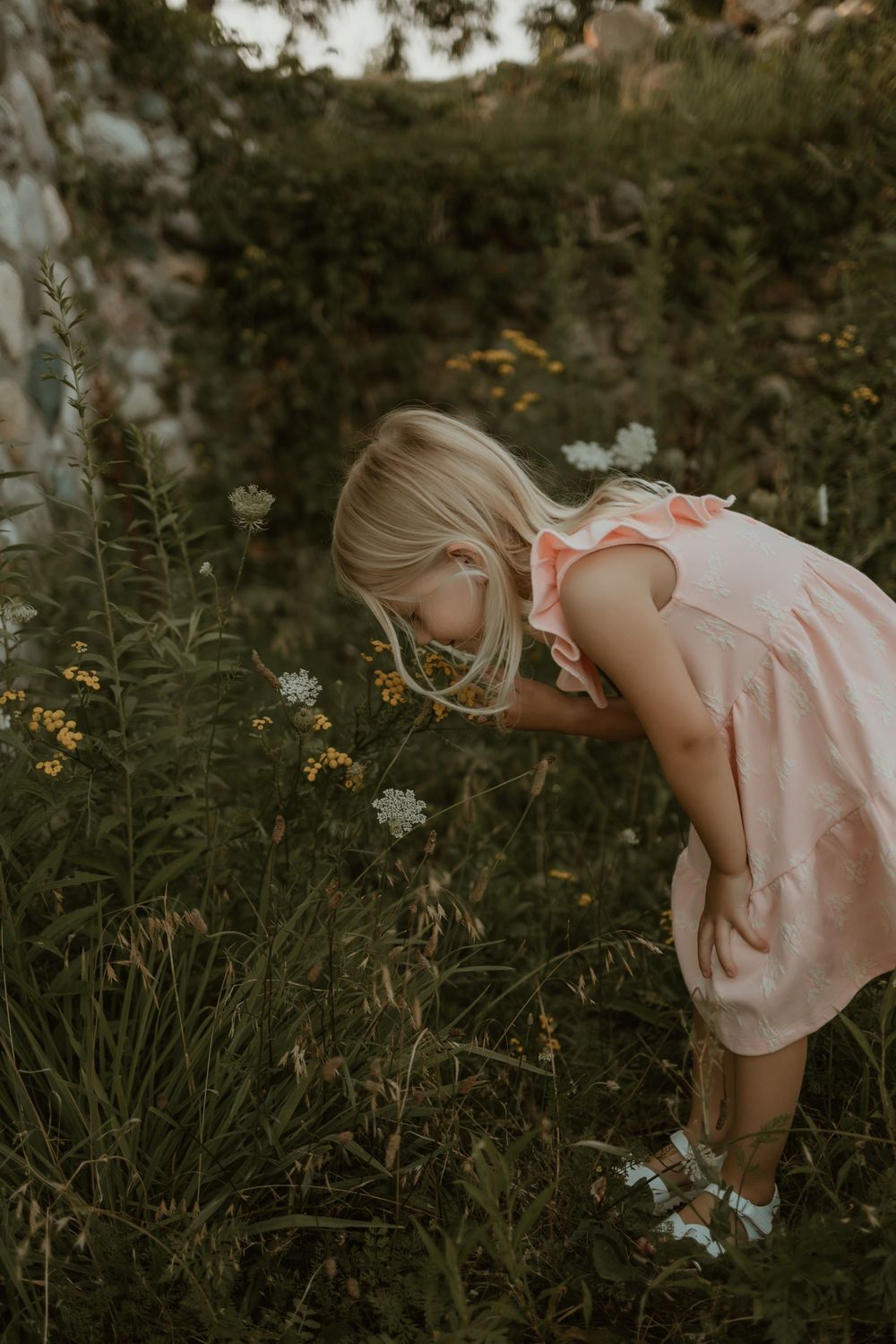 A child in a pink dress explores wildflowers and tall grass in a natural outdoor setting.