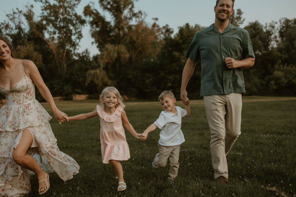 Family holds hands while running together through a grassy field at sunset during a casual outdoor photo session.