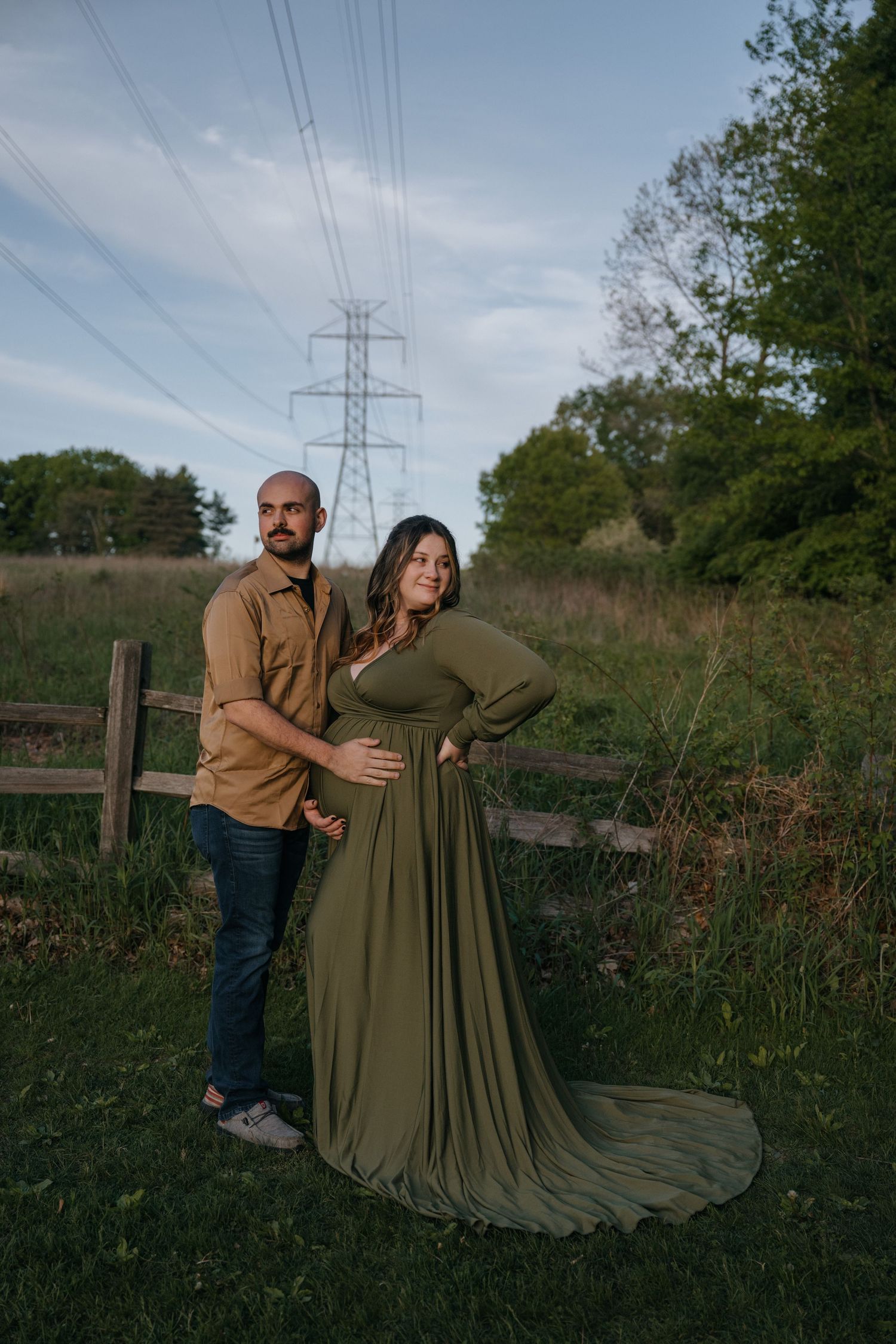 A couple stands together in a field at dusk with power lines and greenery in the background.