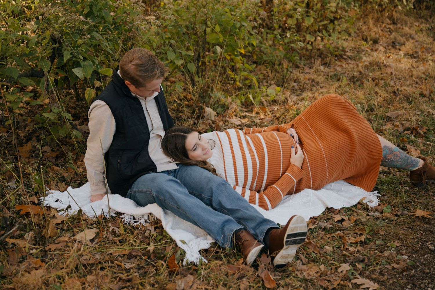Two people relax together on a white blanket outdoors during autumn with fall foliage in the background.