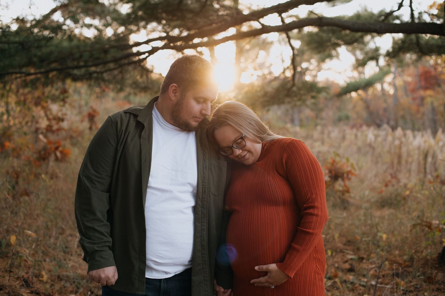 A couple shares an intimate moment at sunset in an autumn field with tall grasses and trees in the background.