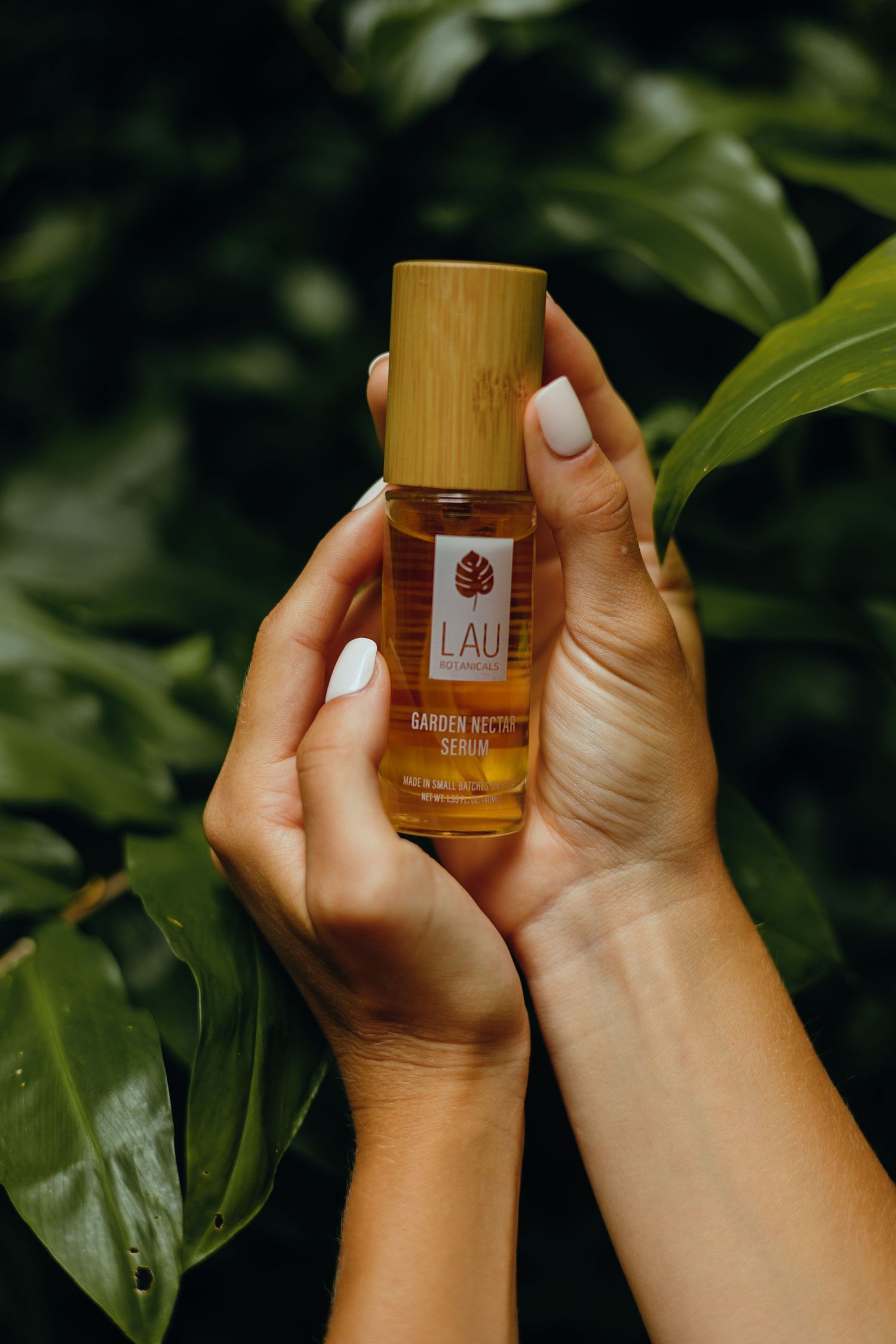 Hands hold a wooden bamboo bottle of beauty product against tropical green leaves.