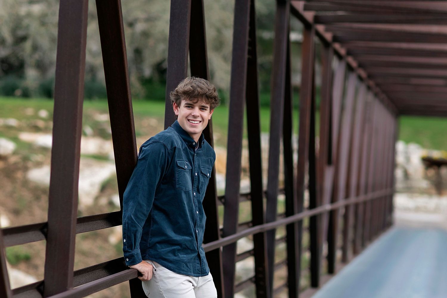Smiling young man in denim shirt leaning on metal bridge railing with green landscape background.