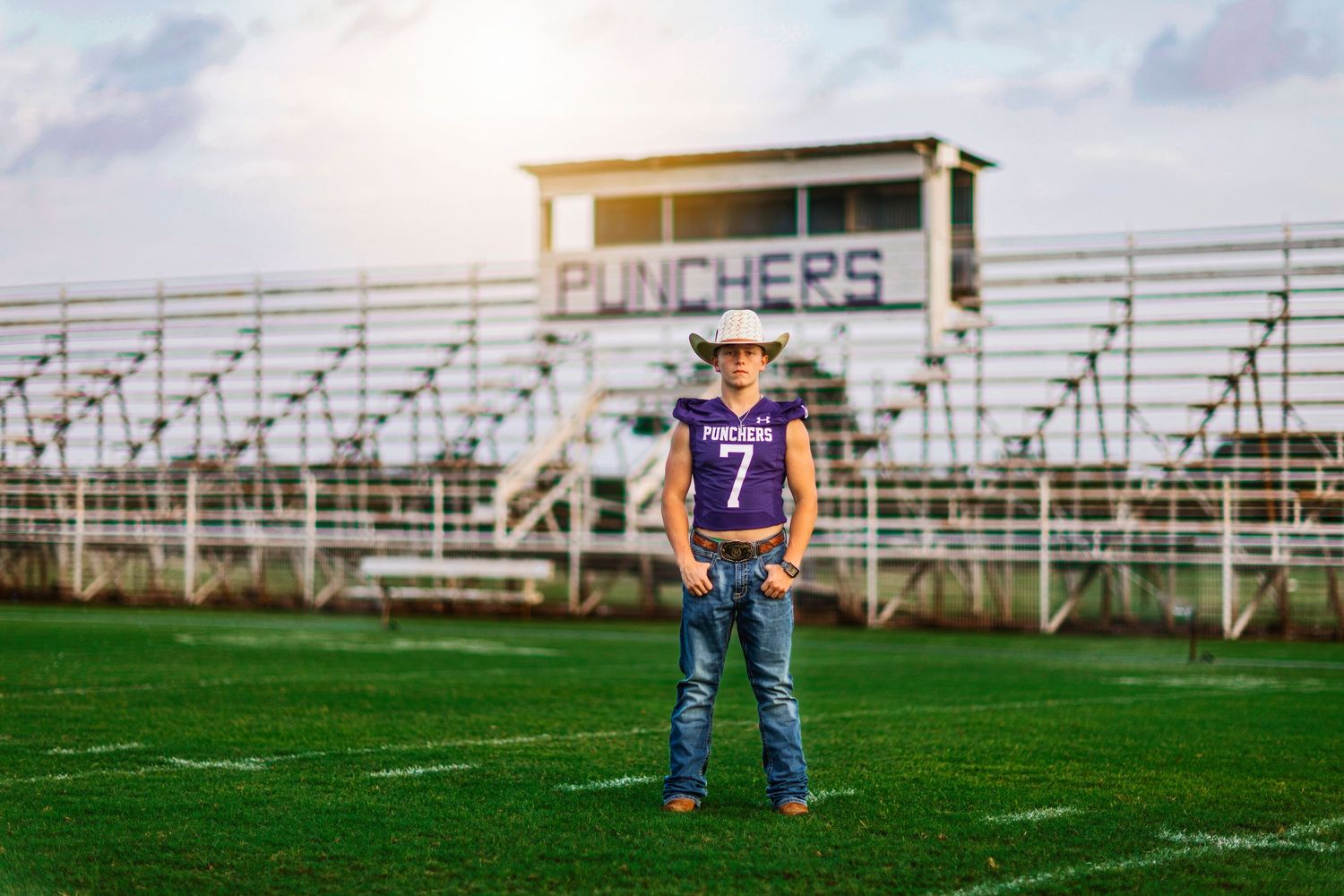 Teen boy in purple Punchers jersey number 7 stands on football field with stadium bleachers behind him.