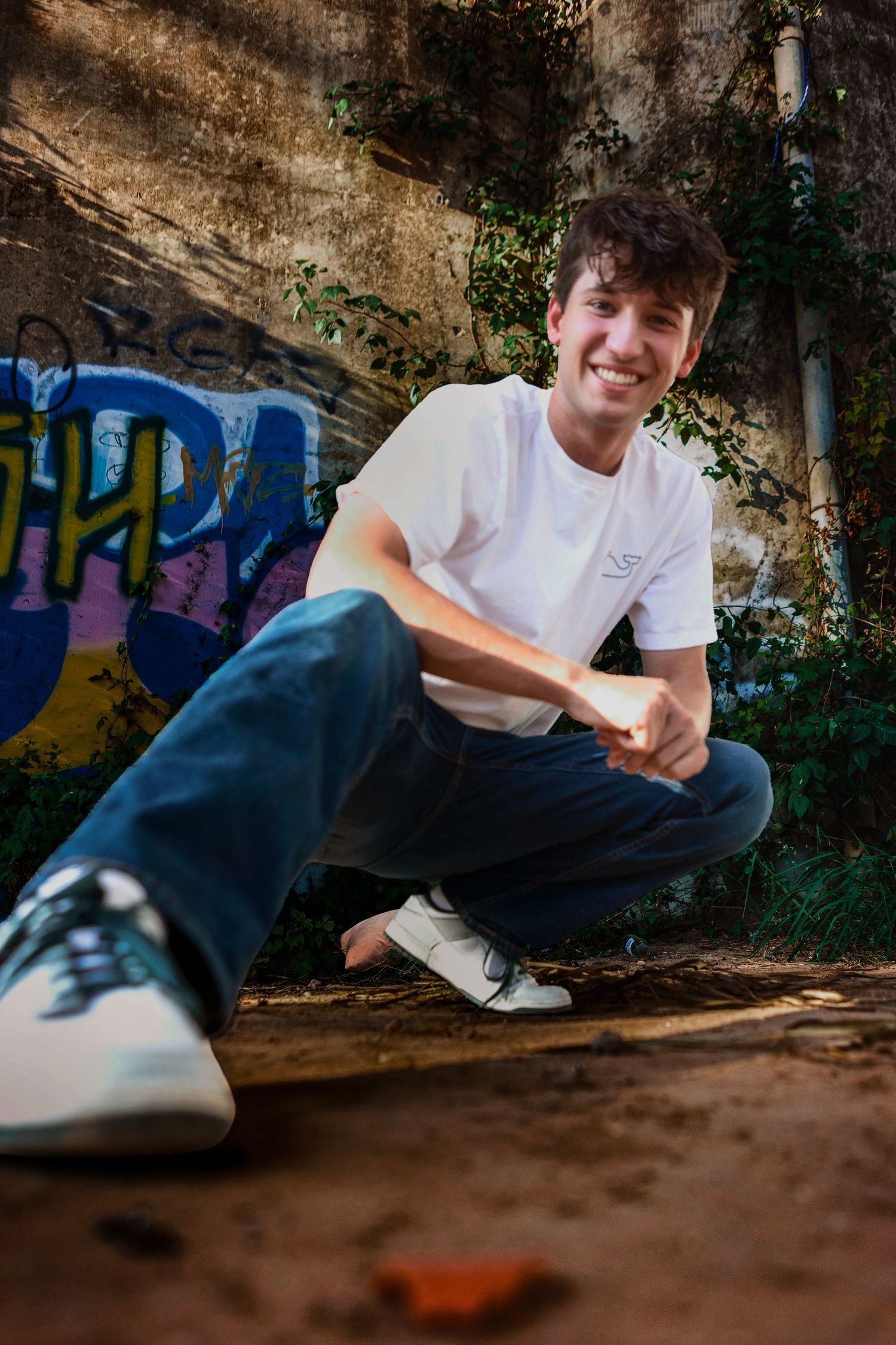 Young man smiling in white tee and jeans, crouching near graffiti wall with greenery in urban outdoor setting.