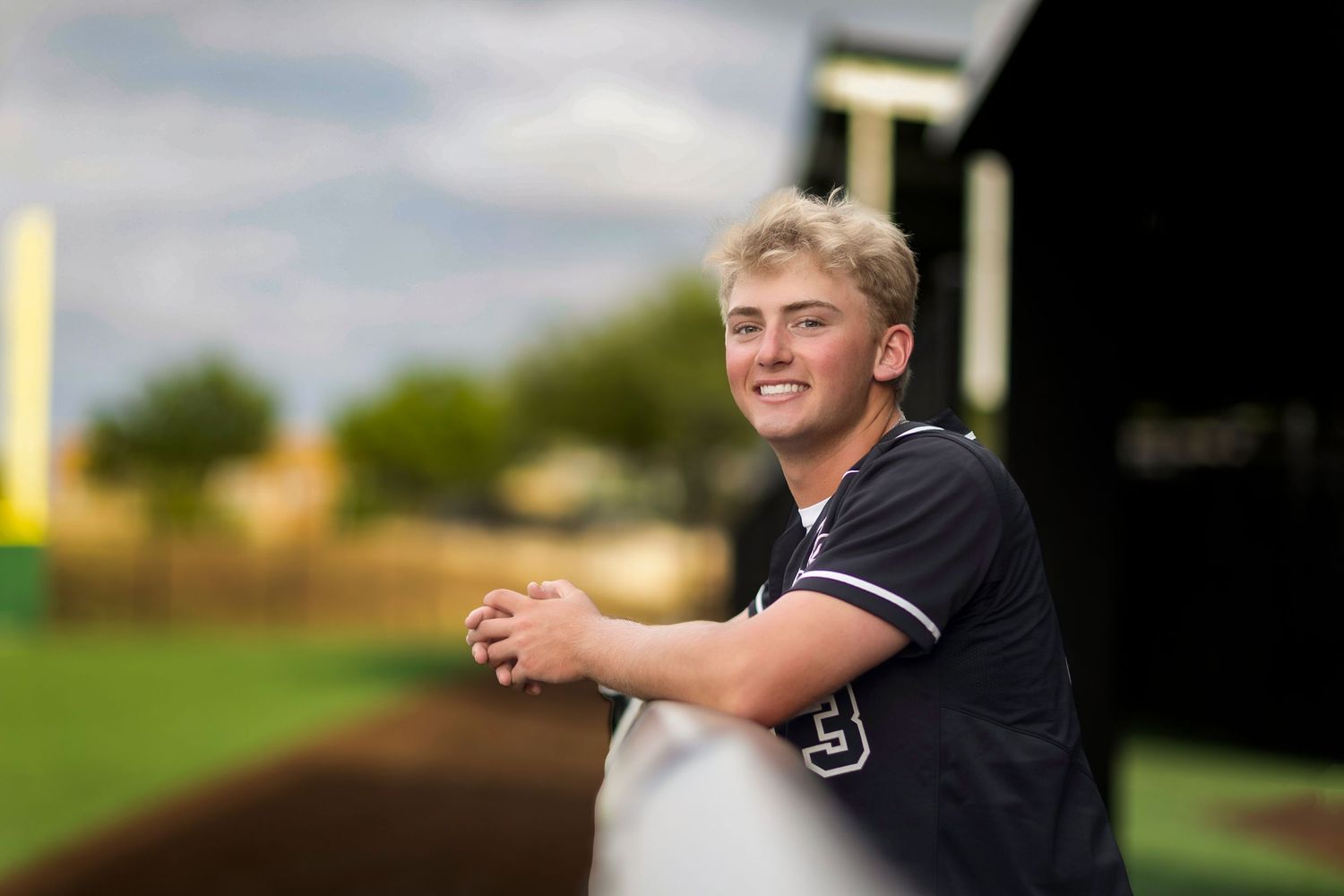 Young teen in baseball jersey leans on dugout railing at baseball field, smiling for senior photos.