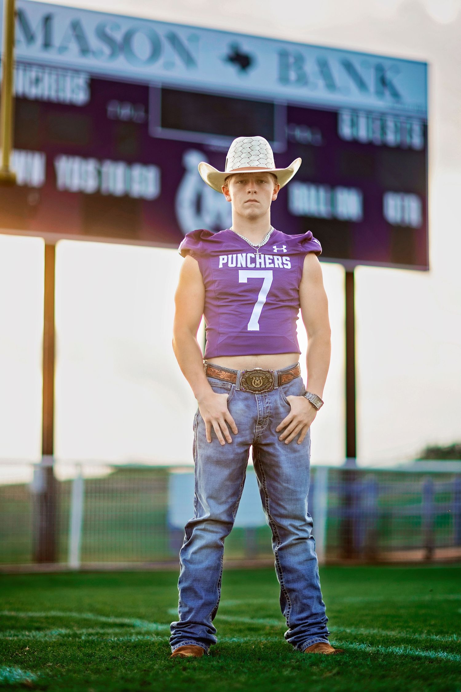 Teen boy in purple Punchers #7 football jersey, cowboy hat, and jeans stands on field near Mason Bank scoreboard.