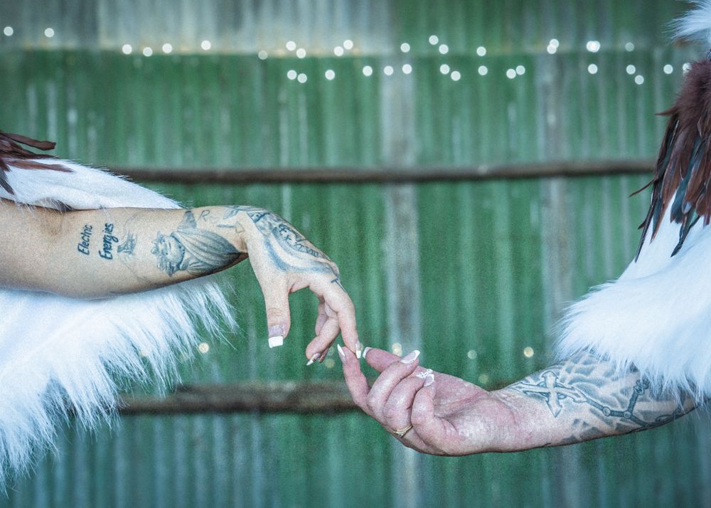 Two tattooed hands reaching towards each other against a green fence background.