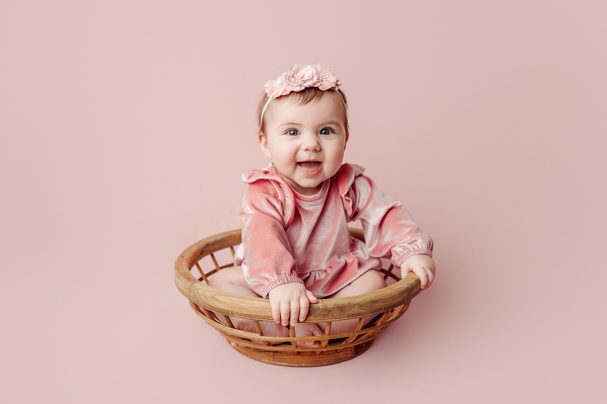 Smiling baby girl in pink outfit and floral headband sitting in a wicker basket against a pink background.