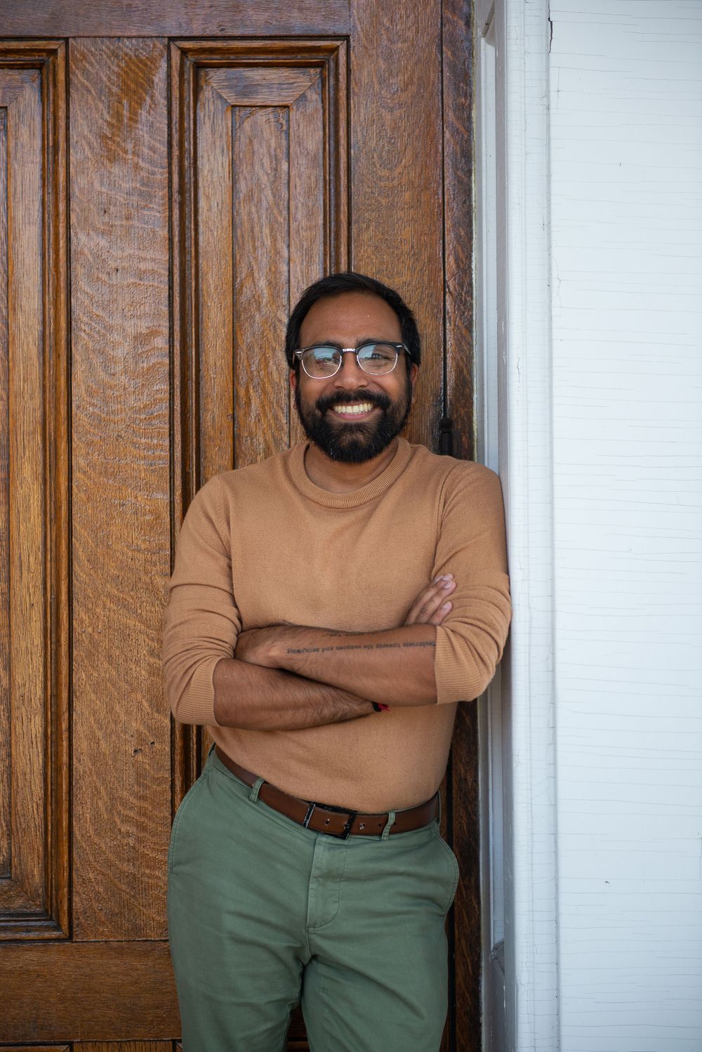 Man in tan sweater and green pants smiles while standing by wooden door.