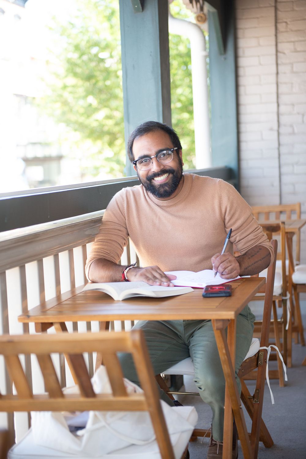 Person writing in notebook while sitting at wooden table on outdoor porch.