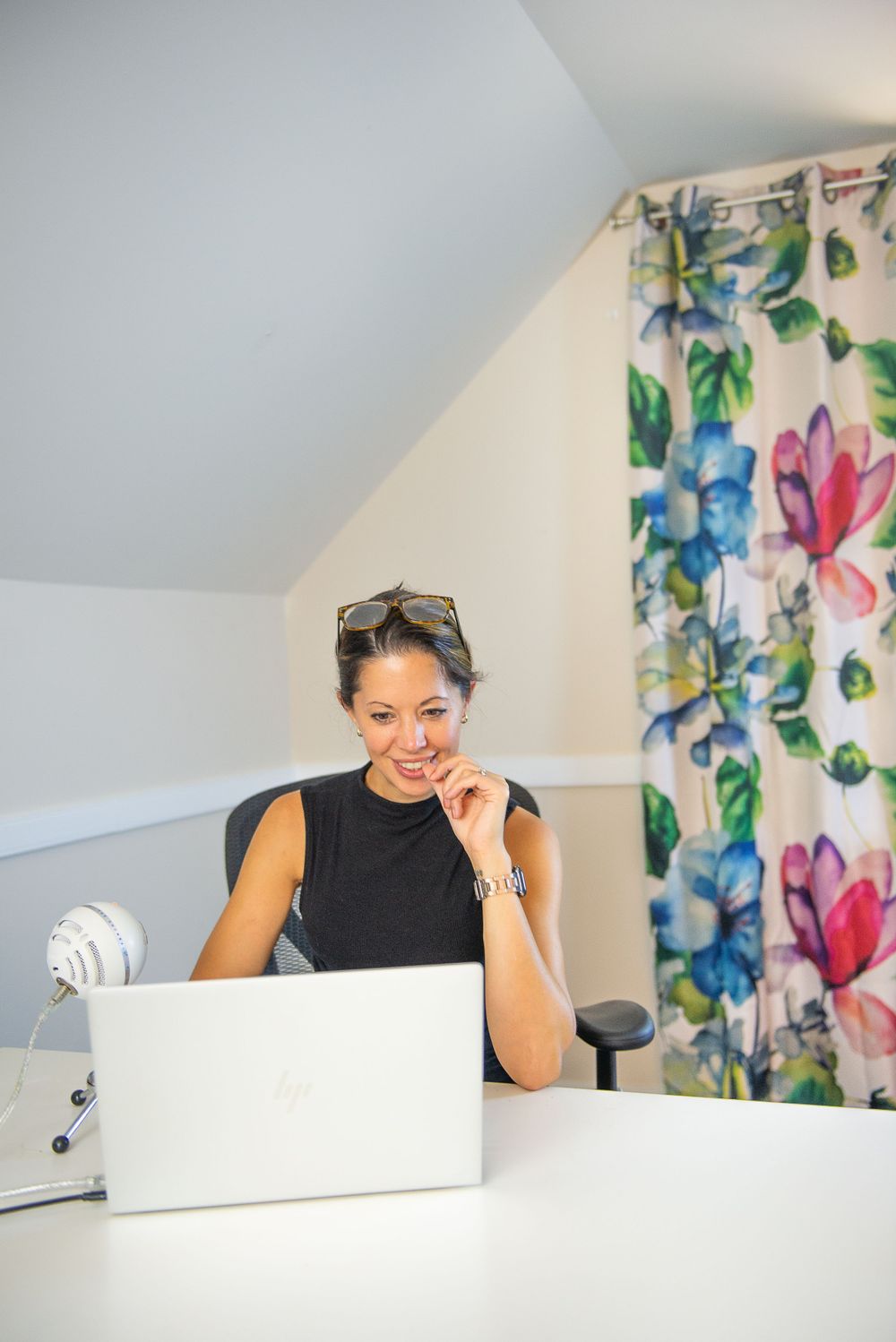A person working on laptop in home office with floral curtains in background.