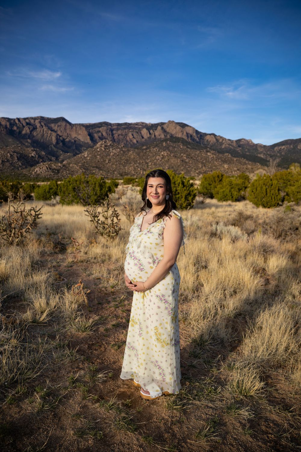 Pregnant woman in floral dress standing in desert landscape with Sandia Mountains backdrop under blue sky.