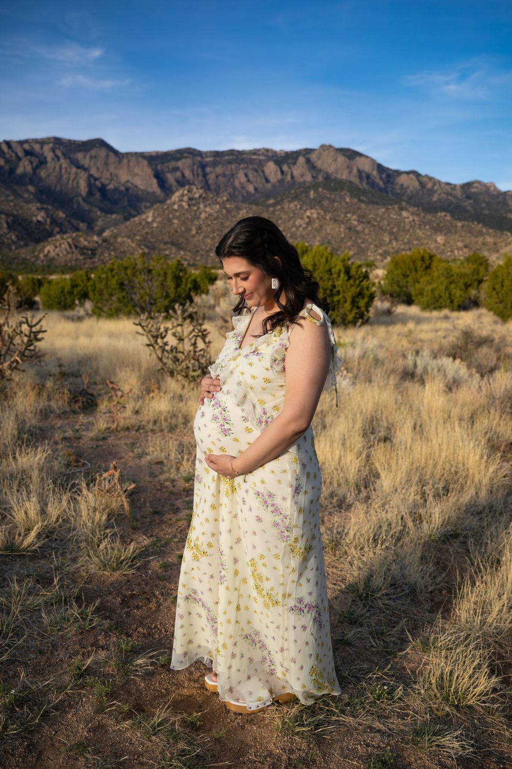 Pregnant woman in floral dress cradling baby bump outdoors with Sandia Mountains backdrop in New Mexico.