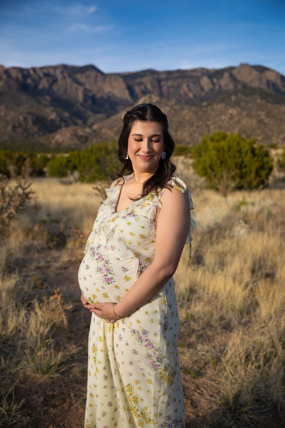 Pregnant woman in floral dress cradling baby bump outdoors with Sandia Mountains in background.
