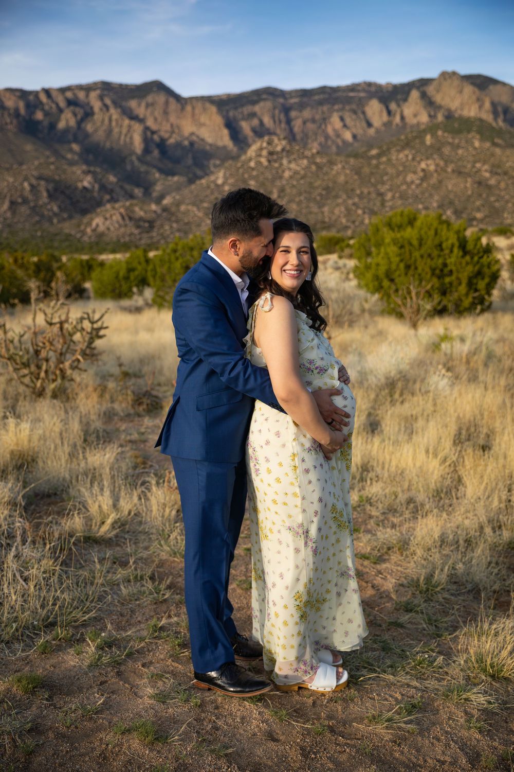 A couple poses in a desert landscape, the man in a blue suit embracing a pregnant woman in a floral dress with mountains behind.