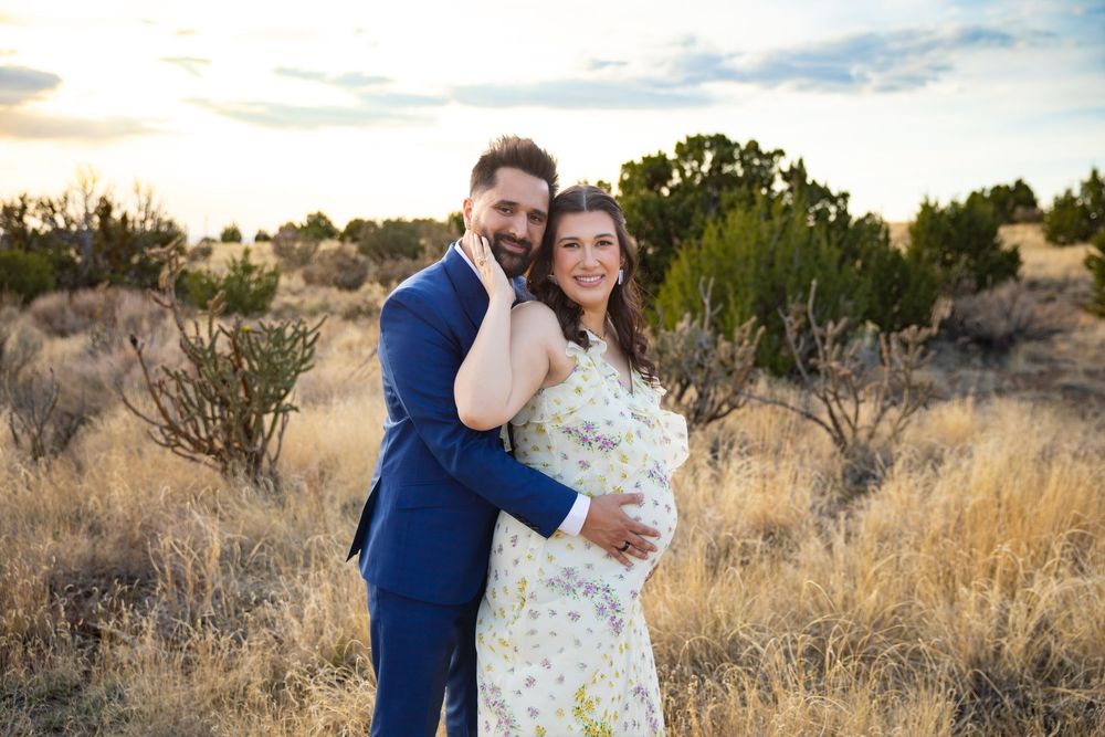 Couple posing in golden field at sunset, man in blue suit embracing pregnant woman in floral dress.