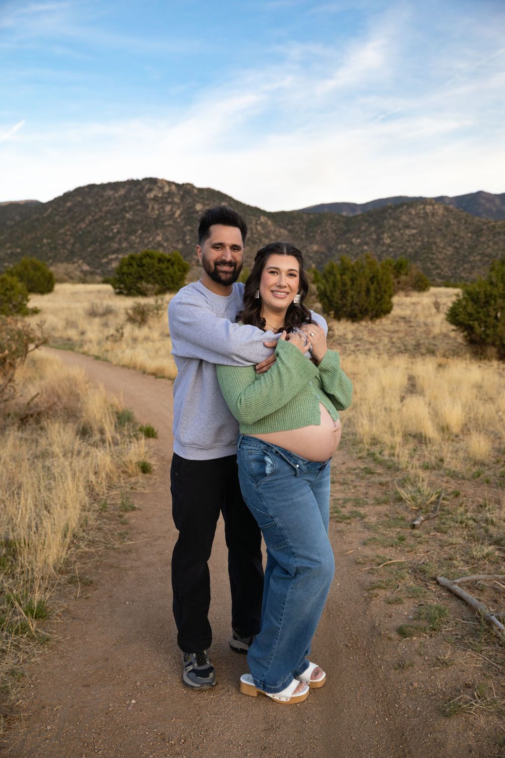 Couple posing on a dirt path in a desert landscape, woman showing baby bump, mountains in background.