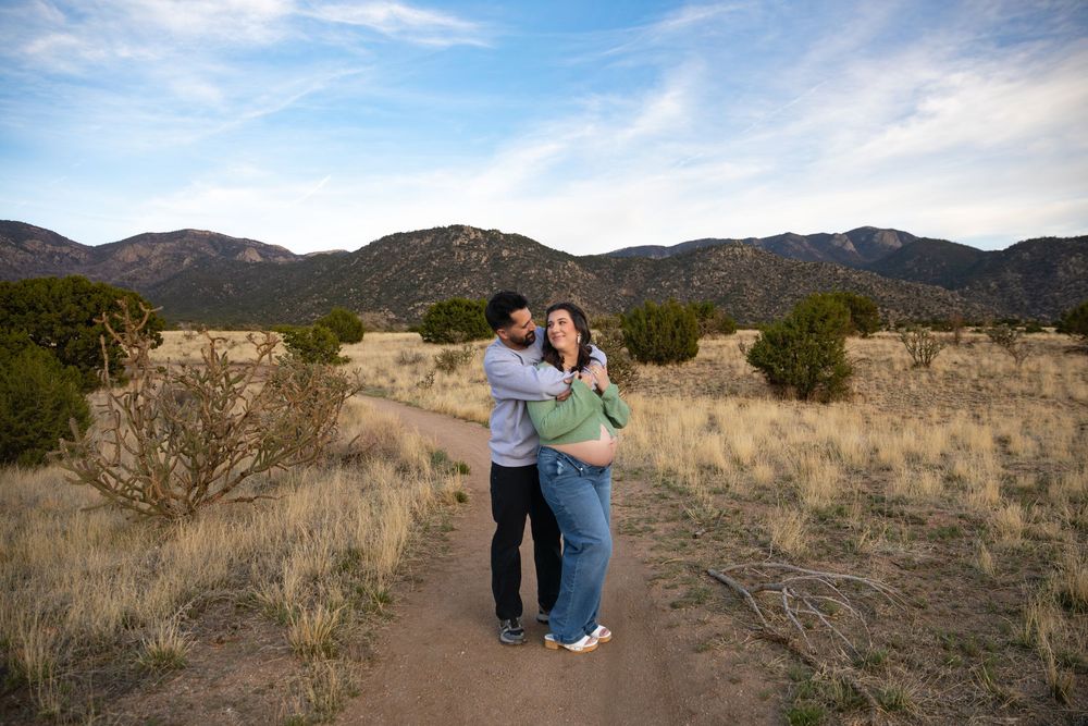 Couple embracing on a desert trail surrounded by golden grasslands and mountain views under a blue sky.