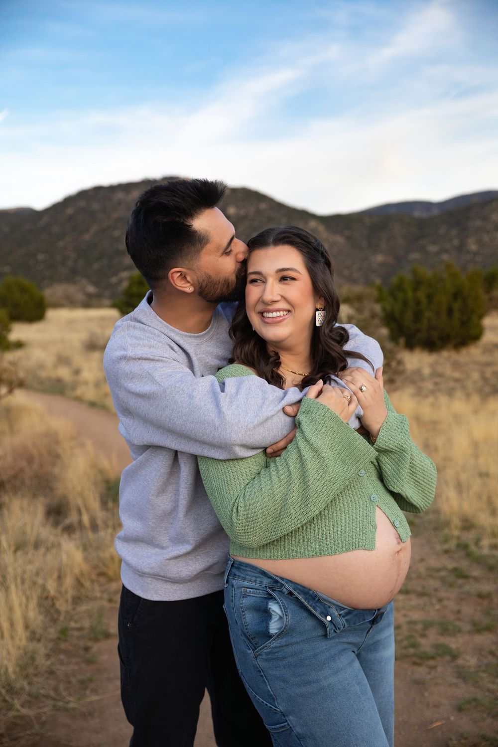 Couple sharing a tender moment outdoors, pregnant woman in green sweater and jeans embraced by partner with mountains behind.
