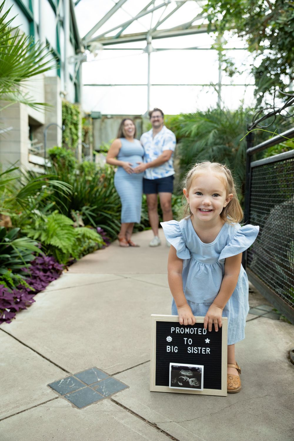 Young girl smiling holding a Promoted to Big Sister sign in a greenhouse with parents in background.