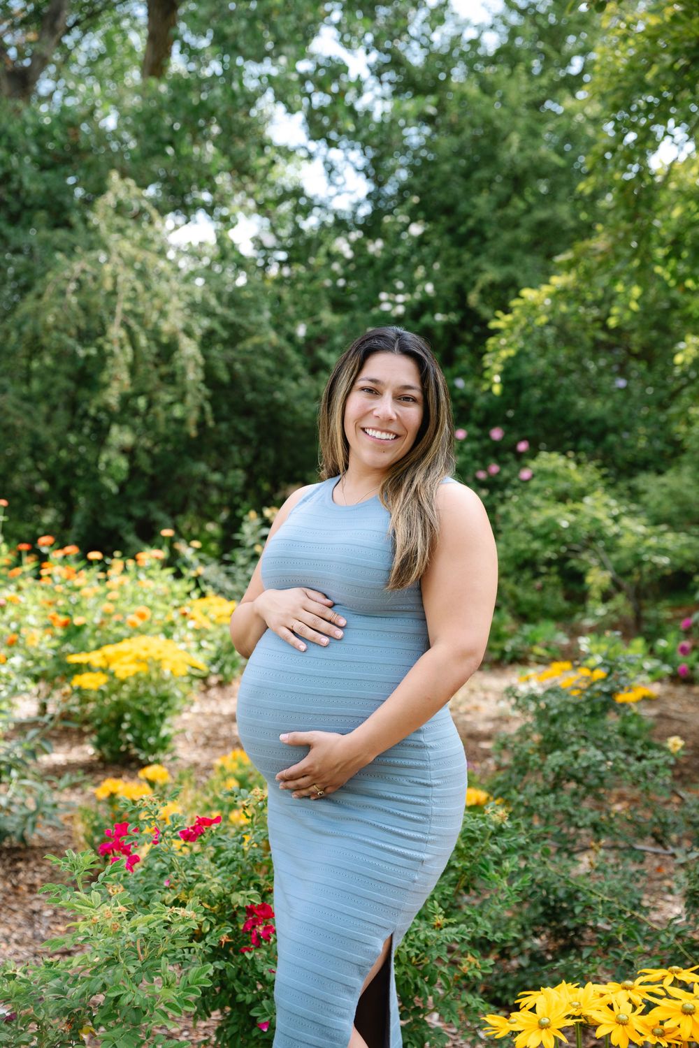 Smiling pregnant woman in blue dress cradling baby bump in colorful garden with yellow and red flowers.