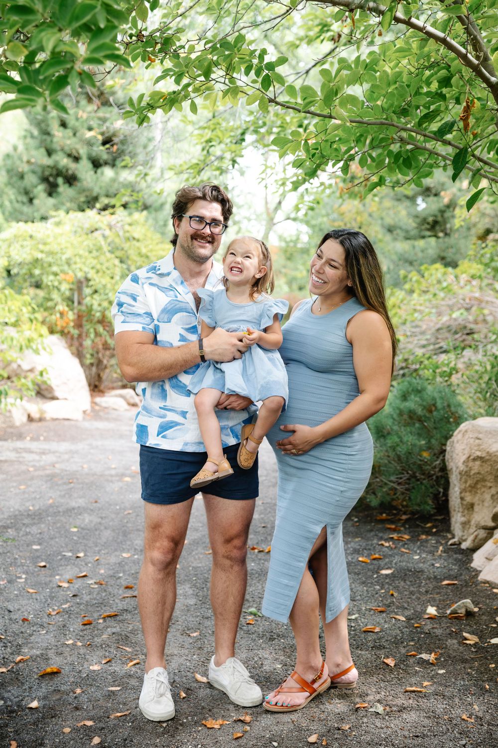 Happy family of three posing outdoors, pregnant mother in blue dress, father holding toddler girl in lush garden setting.