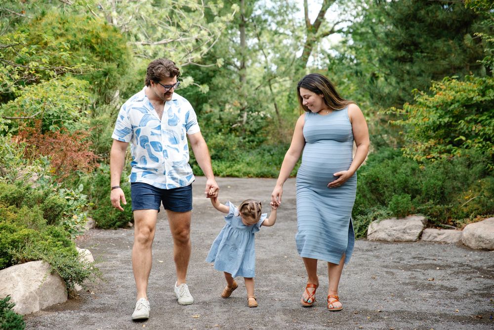 Pregnant woman in blue dress walks with partner and toddler daughter through lush garden path.