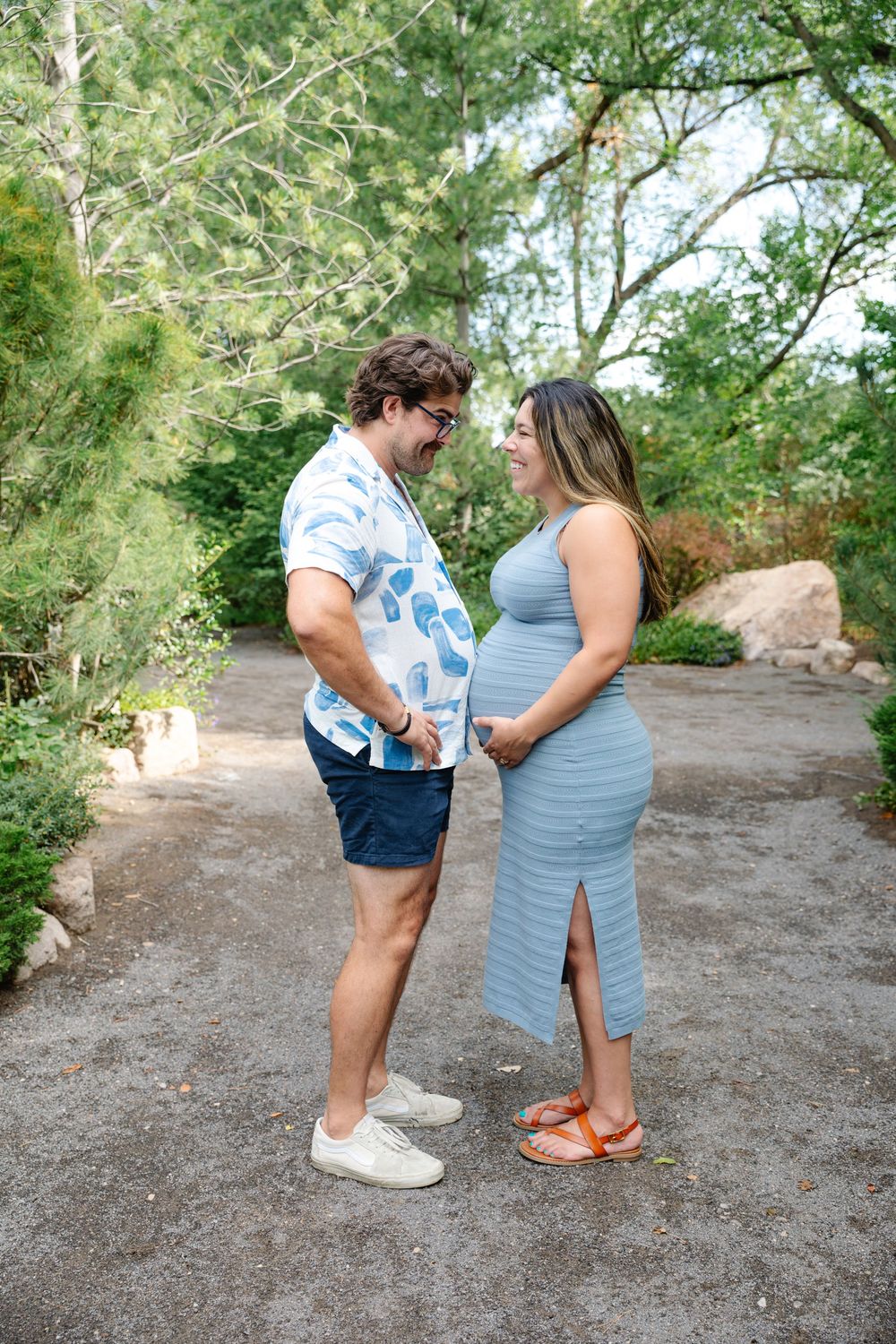 Expecting couple smiling at each other outdoors, woman in blue maternity dress, man in floral shirt, surrounded by lush greenery.