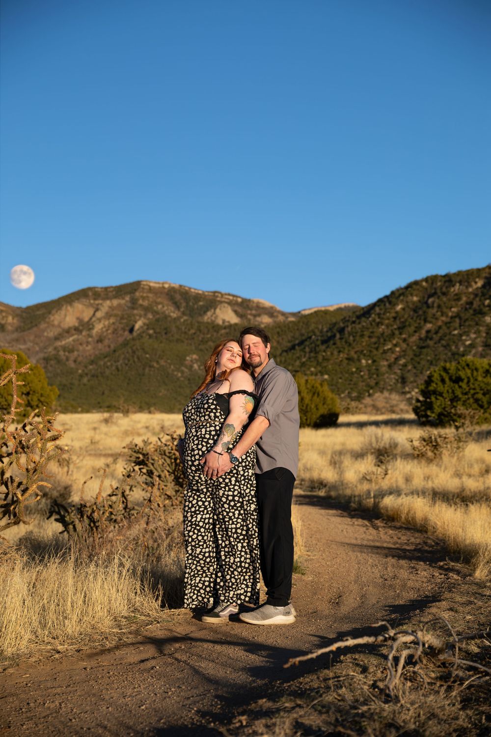 A couple embraces on a desert trail with mountains and a full moon in the background during sunset.