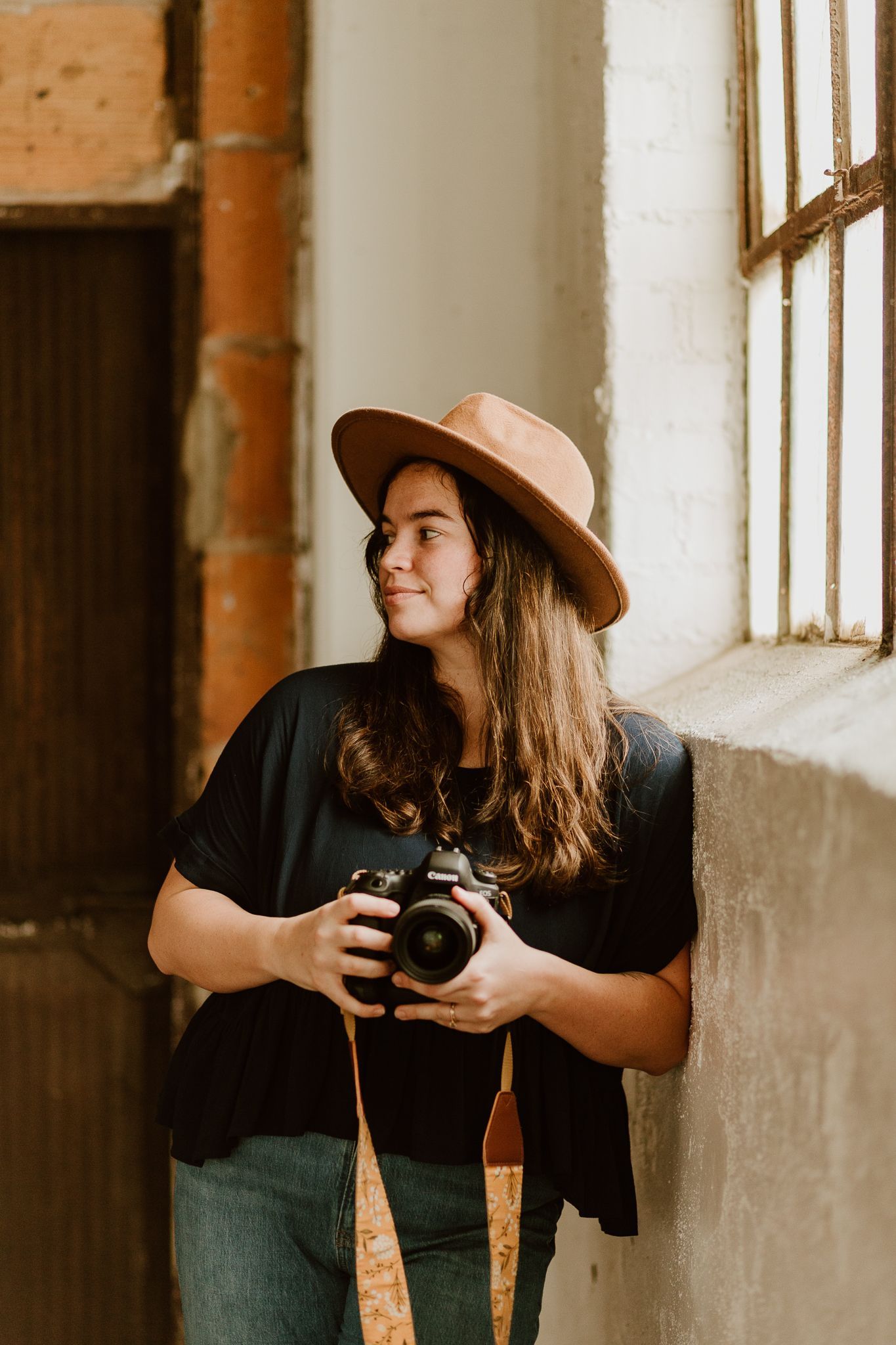 A female photographer in STL wearing a felt hat and holding a camera looks off to the left. She leans on a concrete wall in a loft.