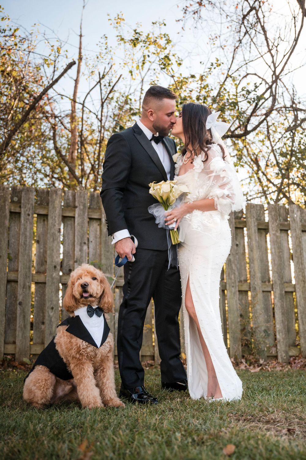 A couple in formal wedding attire poses in a backyard garden with their goldendoodle dog wearing a bow tie.