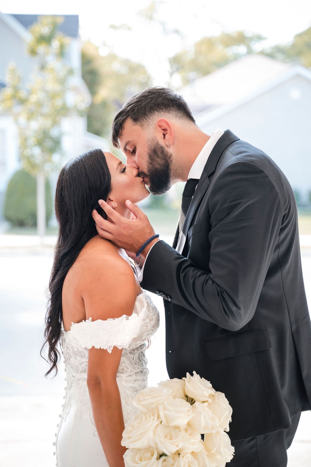 A tender wedding moment captured outdoors as newlyweds share an intimate kiss while holding a white rose bouquet.