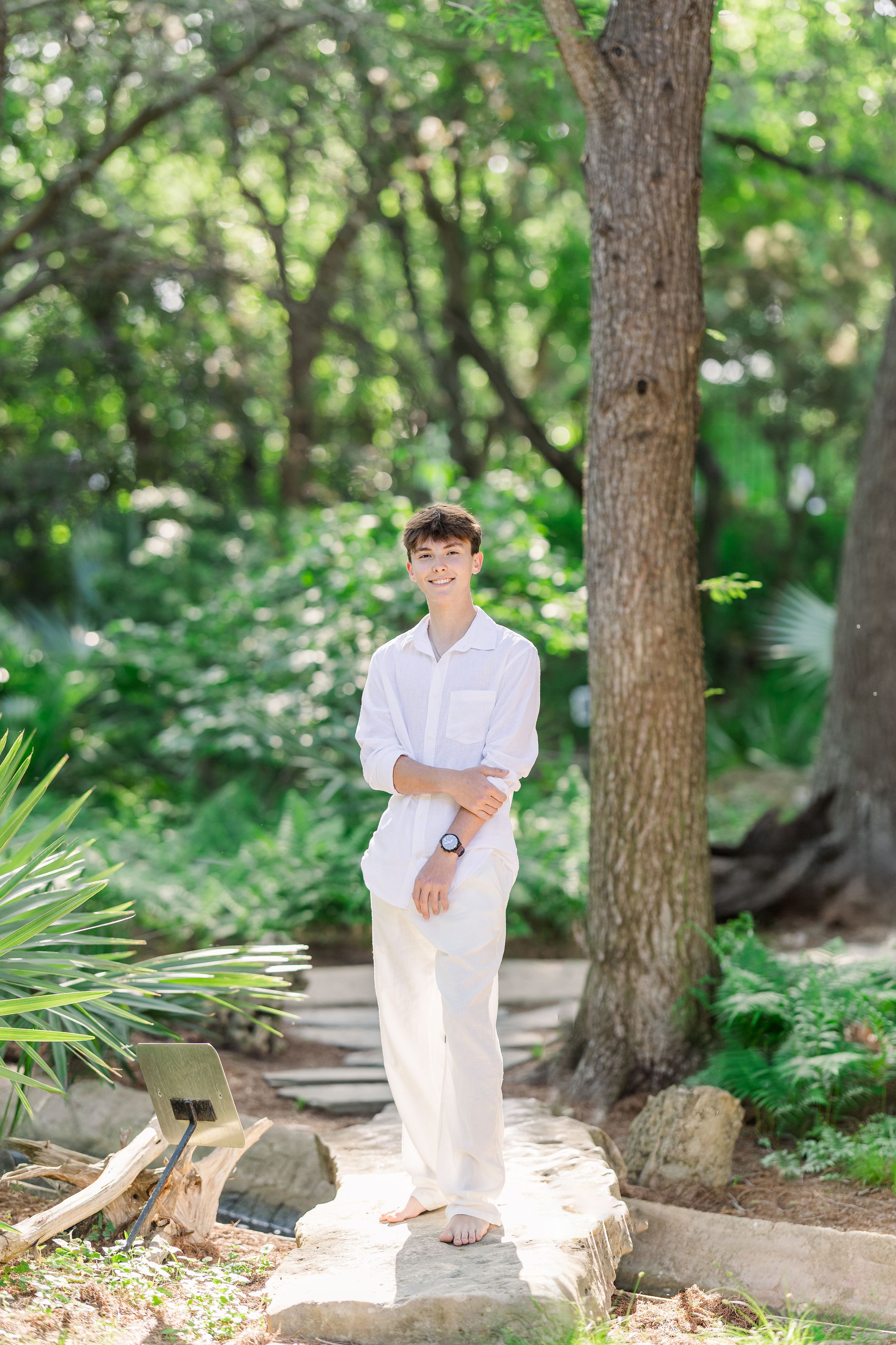 A person in white clothing poses in a forest setting with natural sunlight filtering through the trees.