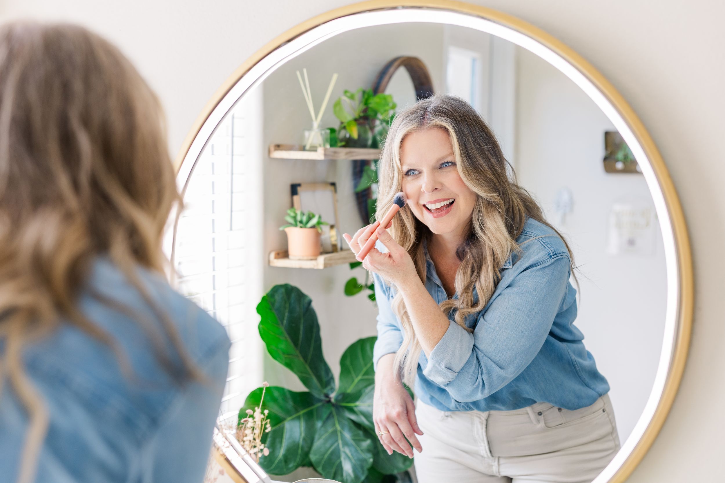 A reflection in a round gold-framed mirror shows a denim shirt and indoor plants in a bright white room.
